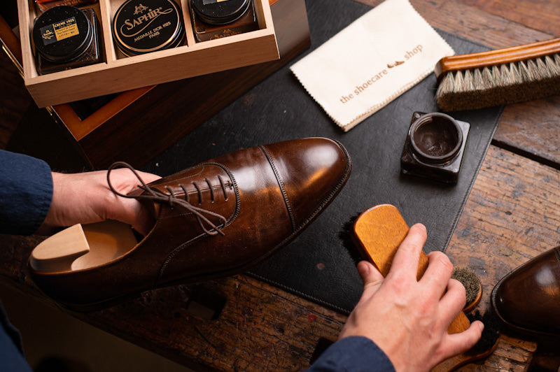 A person polishes a brown leather shoe. A wooden shoe tree is inserted. Shine products, brushes, and a cloth from "the shoecare shop" are present.